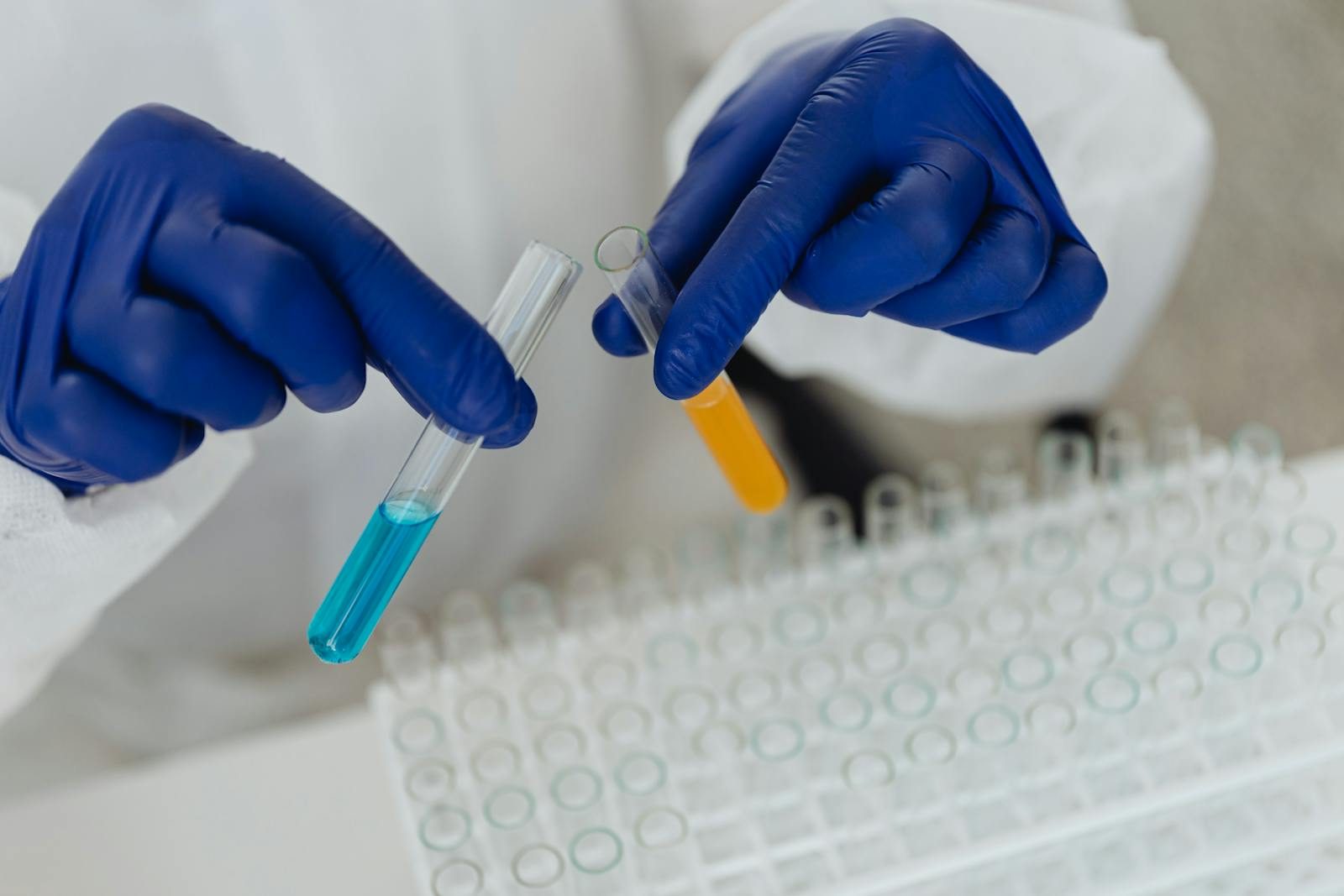 Close-up of lab technician in gloves handling blue and orange liquids in test tubes.